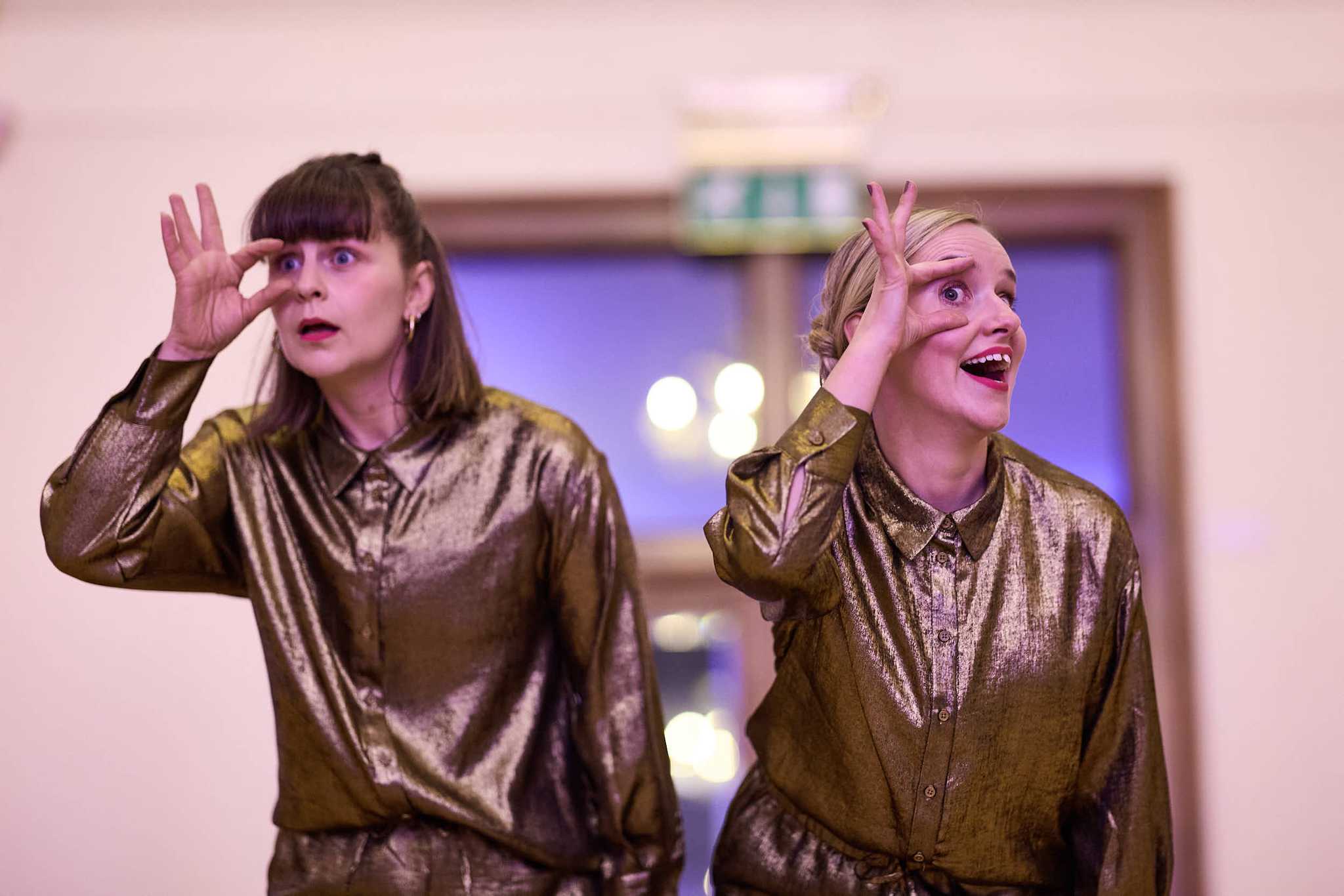 Two female performers dress in gold shiny shirts are staring towards the audience with index fingers and thumbs around ther right eyes, pulling their eyes open, making it a bit intense!